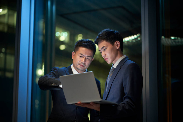 two asian corporate executives standing by the window in office discussing business using laptop computer at night - Photo, Image