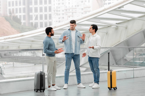 Conflict During Trip. Three Friends Guys Having Quarrel And Misunderstanding Problem During Vacation Travel, Arguing Standing With Suitcases In Modern Airport Terminal Indoors. Full Length Shot - Photo, Image