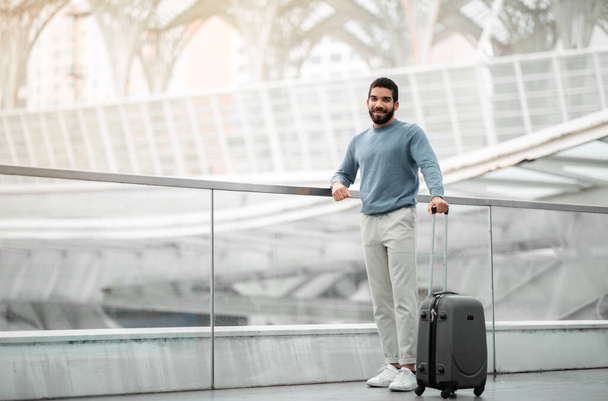 Oferta turystyczna. Pełna długość ujęcia Happy Arabic Tourist Man pozowanie z walizki uśmiechnięty do kamery stojącej na nowoczesnym lotnisku. Pasażer z bagażem czeka na pokład. Wolna przestrzeń - Zdjęcie, obraz