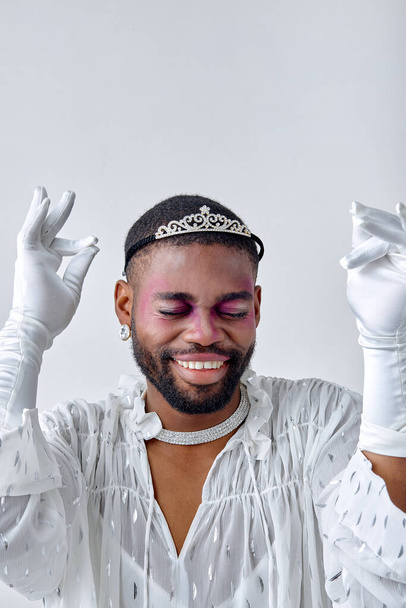 Positive transgender with bright night party makeup in crown dancing isolated over neutral white background. Excited young guy in white shirt and gloves laughing. Lgbtqi, beauty concept - 写真・画像