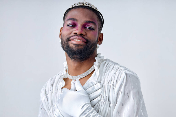 Pleasant and shy 20-25 black bearded man with dark skin of american ethnicity in white sweet shirt and crown. Gay wear fashion style and poses over white studio Background isolated - 写真・画像
