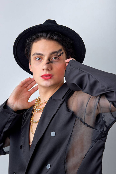 gorgeous american gay man with make up in black fashionable outfit, chain and hat accessories on male, posing isolated on white background studio portrait. People lgbt lifestyle concept. - 写真・画像
