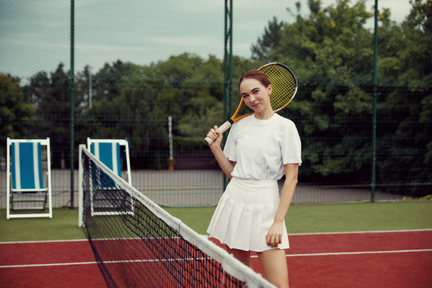 Jovencita alegre de pie mientras sonríe cerca de la red de tenis en la cancha de tenis con raqueta en las manos - Foto, Imagen