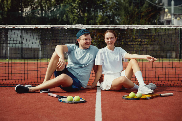 テニスコートの床にラケットとボールで座っている陽気なカップル。友達は笑顔で楽しんでいます。友達とスポーツアクティブゲーム。試合後のテニス選手 - 写真・画像