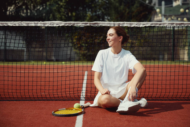 Młoda kobieta z rakietą tenisową w białych ciuchach siedzi na korcie tenisowym w pobliżu siatki i patrzy w prawo uśmiechając się - Zdjęcie, obraz