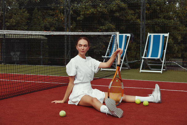 Junge schöne Sportlerin mit Tennisschläger sitzt am Tennisnetz auf dem Tennisplatz. Sportmode. Aktivitäten am Wochenende und Sonntag zur Erholung - Foto, Bild