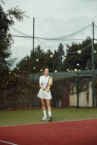 白い衣装を着たテニス選手の縦写真が試合でボールを提供しています。若いです女の子プレイテニスでコート屋外. - 写真・画像