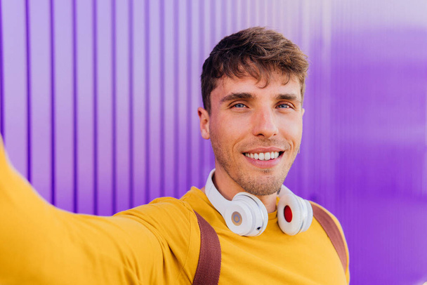 Hombre guapo sonriendo a la cámara sobre un fondo púrpura. Chico feliz aislado tomando una foto selfie con un dispositivo inteligente móvil - Foto, Imagen