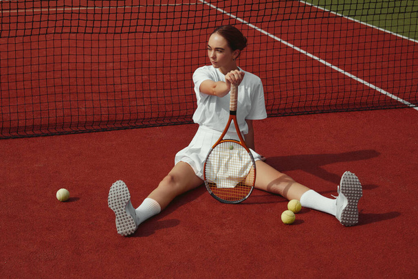 Schöne modische Mädchen sitzt auf dem Platz in der Nähe des Netzes, hält Schläger und trägt weißes Outfit. Sportmode - Foto, Bild