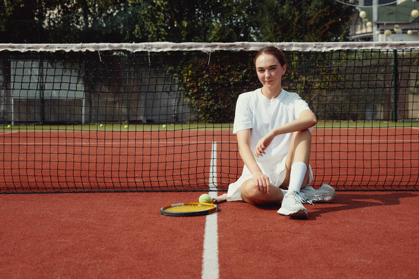 Девушка-спортсмен в белой футболке и платье с ракеткой на теннисном корте сидит у теннисной сетки. - Фото, изображение