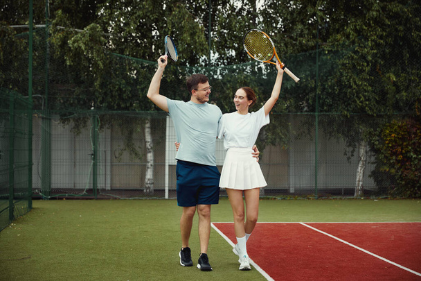 Joyful man and woman are celebrating victory at tennis court. They are laughing and looking at each other while encouraging themselves. Couple is lifting arms with rackets - Photo, Image