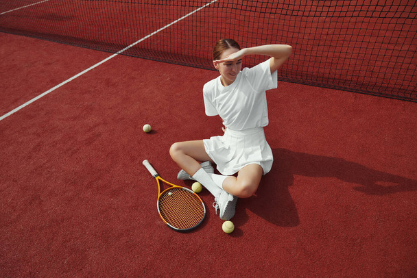 Portrait de la jeune femme en tenue blanche couvrant le visage à la main de la lumière du soleil tout en étant assis sur le sol dans un court de tennis extérieur, prenant une pause de la pratique. - Photo, image