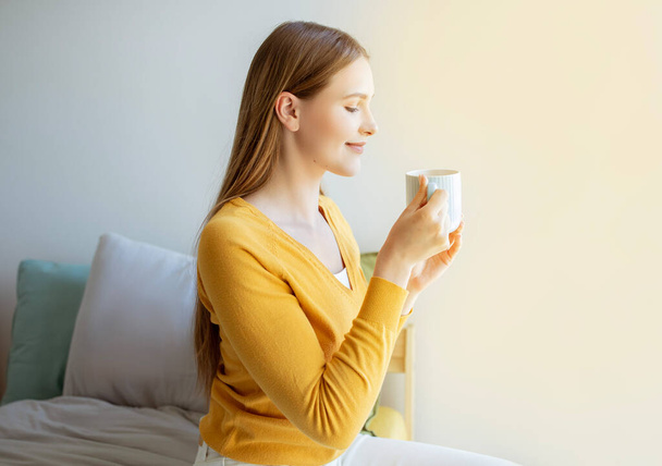 Bonjour, Delight. Heureuse jeune femme odeur arôme de café frais boire tenant tasse assis sur le lit dans sa chambre à la maison. Vue latérale de dame joyeuse bénéficiant d'une boisson chaude dans une tasse - Photo, image