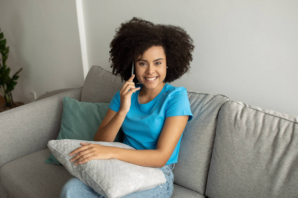 Καλά νέα, κουτσομπολιό. Ευτυχής όμορφη νεαρή μαύρη σγουρή γυναίκα καλώντας από το smartphone, μιλώντας με φίλο στον καναπέ στο εσωτερικό του σαλονιού. Ελεύθερος χρόνος, επικοινωνία στο σπίτι - Φωτογραφία, εικόνα