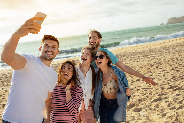 Vrolijke duizendjarige Europese en Arabische vrienden hebben plezier, selfie nemen op smartphone, genieten van vrije tijd op het strand van de oceaan. App voor blog en sociaal netwerk, outdoor party, vakantie en reis - Foto, afbeelding
