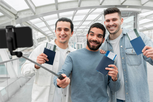 Share Your Travel Moments. Group Of Cheerful Travelers Guys Making Selfie For Fun, Posing With Passports And Boarding Passes Tickets In Modern Airport. Technology And Transportation - Foto, imagen