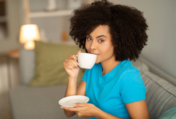 Улюблений свіжий смачний напій. Усміхнена молода чорна кучерява жінка п'є чашку гарячої кави на дивані, насолоджується вільним часом, відпочиває наодинці ввечері в інтер'єрі вітальні. Чай і відпочинок вдома
 - Фото, зображення