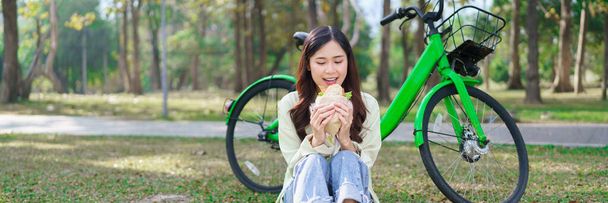 Γυναίκες που τρώνε σάντουιτς με ευτυχία ενώ κάθονται στο γρασίδι για να ξεκουραστούν μετά την ποδηλασία στο πάρκο. - Φωτογραφία, εικόνα
