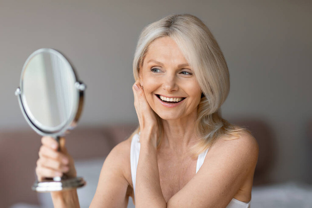 Пожилая концепция природной красоты. Счастливая пожилая женщина смотрит в зеркало, трогает лицо и улыбается в отражении, наслаждаясь новыми продуктами по уходу за кожей - Фото, изображение