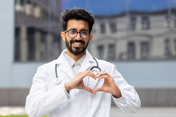 Portrait of friendly doctor, Hindu man in white medical coat smiling and looking at camera, showing heart with hands gesture of kindness. - Photo, Image