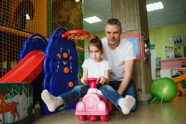 Ένα χαρούμενο κορίτσι με τον πατέρα της παίζει σε ένα παιδικό κέντρο, ένα κορίτσι οδηγεί μια γραφομηχανή.. - Φωτογραφία, εικόνα