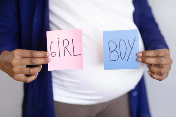 Millennial african american pregnant lady with big belly shows boy or girl paper on gray wall background, cropped. Expectation of child, health care, gender definition, party, ad and offer - Foto, imagen