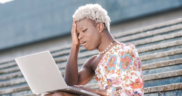 Afrikaanse jonge transgender vrouw zitten op de buitentrap, met behulp van een laptop en glimlachen op een zonnige dag - Foto, afbeelding