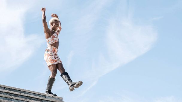 Niedriger Winkel einer unbeschwerten jungen afroamerikanischen Transgender-Person in buntem Kleid, die vor blauem Himmel tanzt und lächelt, während sie an sonnigen Tagen die Freiheit genießt - Foto, Bild