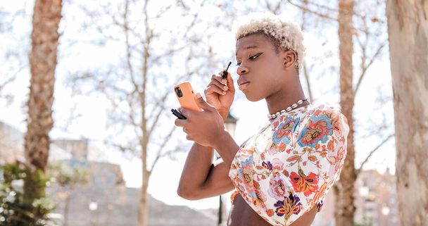 Concentrado joven afroamericano transexual en vestido colorido con pelo rizado y collar aplicando rimel en las pestañas mientras mira la pantalla del teléfono inteligente en la calle - Foto, Imagen