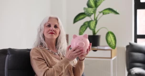 Mulher sênior sorrindo sacode banco porquinho sentado em poltrona escura. Pessoa feminina segura caixa de dinheiro rosa nas mãos. Conceito de acumulação de dinheiro câmera lenta - Filmagem, Vídeo