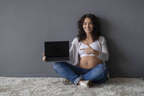 Pregnancy Blog. Smiling Expectant Female Pointing At Laptop With Blank Black Screen While Sitting On Floor Against Grey Wall Background, Happy Pregnant Woman Advertising Website, Mockup - 写真・画像