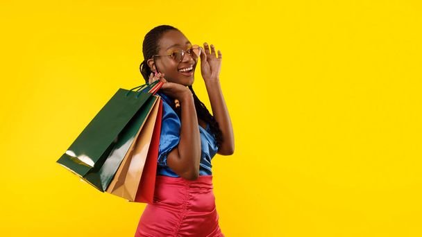 Summer Shopping And Sales. Cheerful Black Lady With Paper Shopper Bags Smiling To Camera, Posing Wearing Sunglasses And Bright Clothes Near Free Space For Text Over Yellow Background, Panorama - Photo, Image