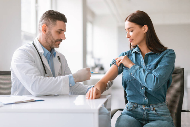 Inoculation time. Doctor man wearing gloves and making vaccination shot to young female patient, inserting syringe to her arm, woman getting vaccine shot against viruses in modern clinic - Foto, Imagen