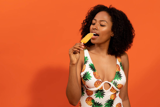 Summer mood concept. Happy hot sexy attractive young black woman wearing trendy colorful swimsuit having delicious ice pop, chilling on orange studio background, copy space - Photo, Image