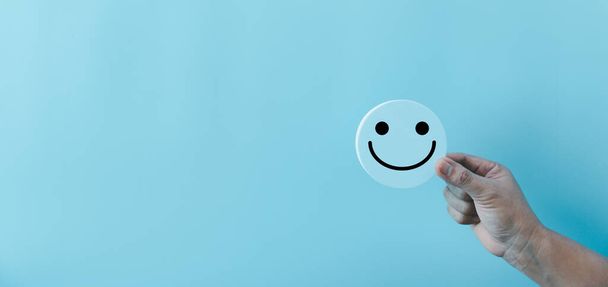 Žena ruka drží šťastný obličej úsměv tvář ikona na kulaté modré objekt. Zkušenosti zákazníků a služby s konceptem spokojenosti. pozitivní myšlení, hodnocení duševního zdraví, světový den duševního zdraví. - Fotografie, Obrázek