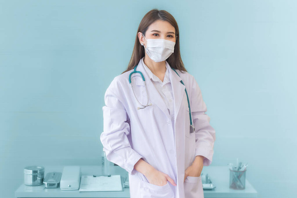 Professional young Asian woman doctor standing with pickpocket wearing a white robe wearing a mask and stethoscope. Coronavirus protection prevent disease and health concept. - Photo, Image