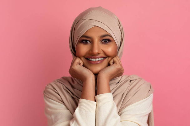 Przyjemne wieści. Marzycielska młoda muzułmanka w hidżabie spoczywająca na rękach i patrząca w kamerę, uśmiechnięta muzułmanka ubrana w headscard pozująca na tle różowego studia, kopiująca przestrzeń - Zdjęcie, obraz