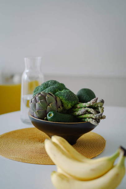 Gesunde Ernährung. Frisches Gemüse auf dem Teller und Bananen auf dem Tisch in der Küche. Leuchtend grünes Gemüse für ein gesundes Leben. Artischocke, Avocado, Spargel, Brokkoli und Bananen aus nächster Nähe - Foto, Bild