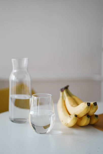 Bright healthy bananas and clean water for the breakfast. The glass with fresh water on the table with ripe fruits. Healthy lifestyle. Bright yellow kitchen in morning light.  - Photo, image