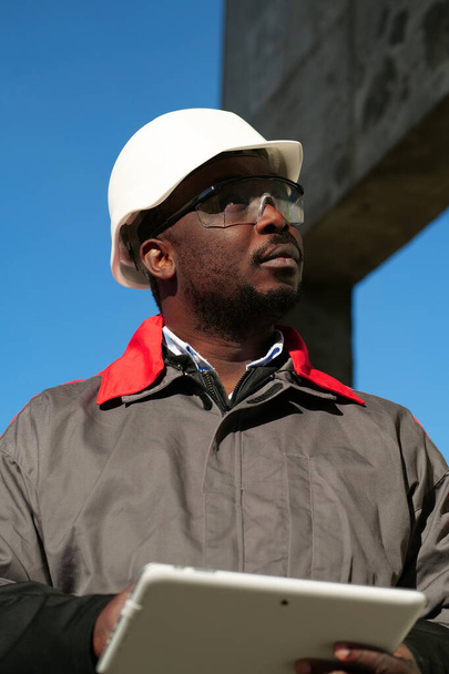 Afrikkalainen amerikkalainen työntekijä kypärä ja suojaava suojalasit tabletti tietokone seisoo rakennustyömaalla - Valokuva, kuva