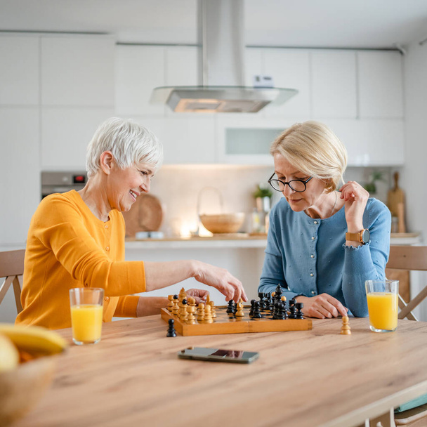 due anziane donne mature caucasiche donne amiche o sorelle giocano a scacchi per il tempo libero gioco da tavolo a casa divertirsi trascorrere del tempo insieme a casa luminosa foto copia spazio - Foto, immagini