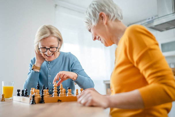 duas mulheres maduras seniores caucasianas mulheres amigas ou irmãs jogar jogo de tabuleiro de xadrez de lazer em casa se divertir passar o tempo juntos em casa brilhante espaço de cópia da foto - Foto, Imagem