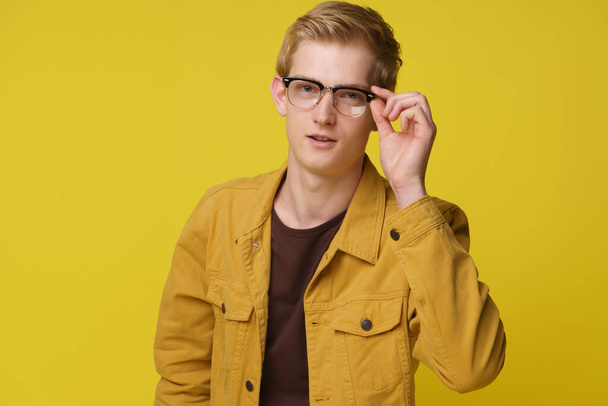 Όμορφος νέος 20άρης με γυαλιά και κίτρινο μπουφάν απομονωμένο φόντο στούντιο. Άντρας κρατώντας γυαλιά στο πρόσωπό του με το χέρι - Φωτογραφία, εικόνα