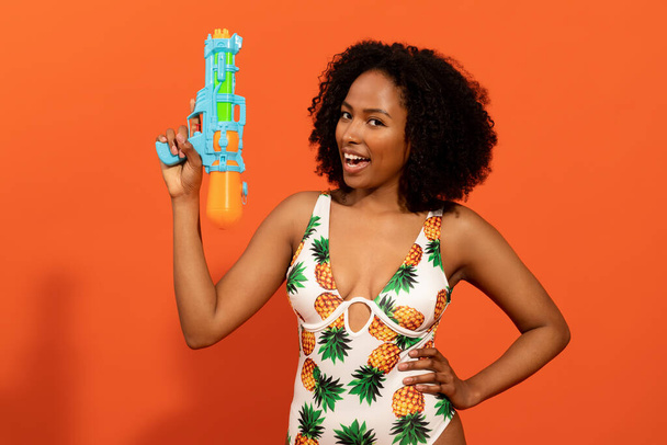 Playful pretty young black woman in colorful swimsuit holding water gun, having fun on orange studio background, copy space. Equipment for water activity in swimming pool while summer vacation - Fotoğraf, Görsel