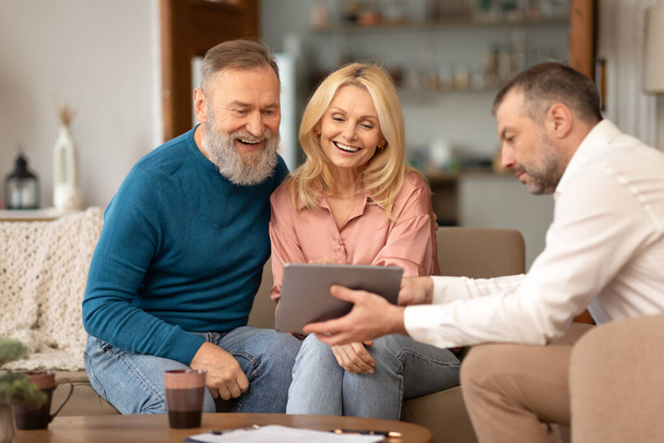 Professional Agent Showing Tablet Computer To Excited Senior Couple Offering Digital Services Sitting On Couch At Home. Real Estate Buyers Choosing House Property With Specialist. Selective Focus - Photo, Image