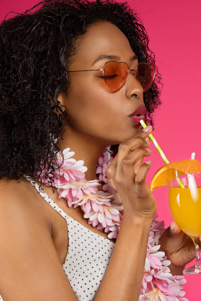 Señora rizada afroamericana milenaria satisfecha en gafas de sol bebiendo cóctel, disfrutar de vacaciones tropicales aisladas sobre fondo rosa, estudio, vertical. Diversión de verano, relax y vacaciones - Foto, Imagen