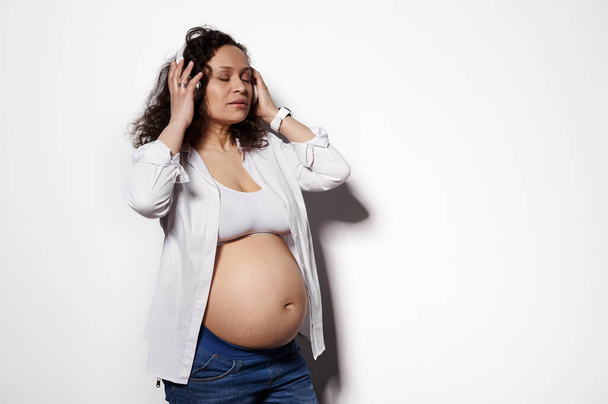 Photo studio portrait of delightful pregnant woman showing her belly, wearing wireless headphones, isolated on white background. People. Lifestyles. Pregnancy. Maternity. Leisure activity. Copy space - Foto, imagen