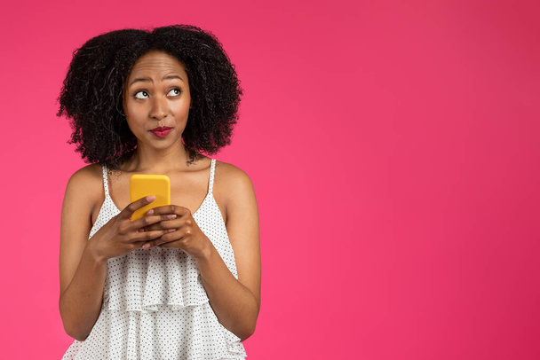Düşünceli neşeli kıvırcık milenyum Afrikalı Amerikalı kadın telefonuyla pembe arka planda, stüdyoda izole edilmiş boş bir yere bakıyor. İyi haber mesajı, satış ve blog, teklif ve reklam - Fotoğraf, Görsel