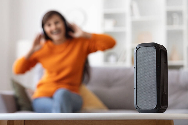 Selective focus on modern wireless musical column standing on table while unrecognizable woman listening to music, enjoying favorite song, sitting on sofa at home. Domestic leisure and gadgets - Фото, зображення