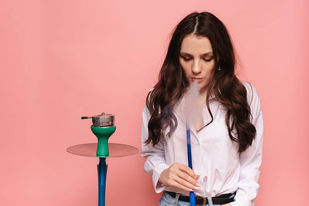 Young, attractive woman smokes hookah, sheesha on a pink background. The pleasure of smoking. - Foto, immagini